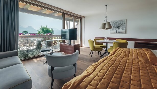 Une chambre d’hôtel comprenant un lit, une télévision, un bureau avec des chaises, un balcon avec vue sur les montagnes et un tableau au mur.