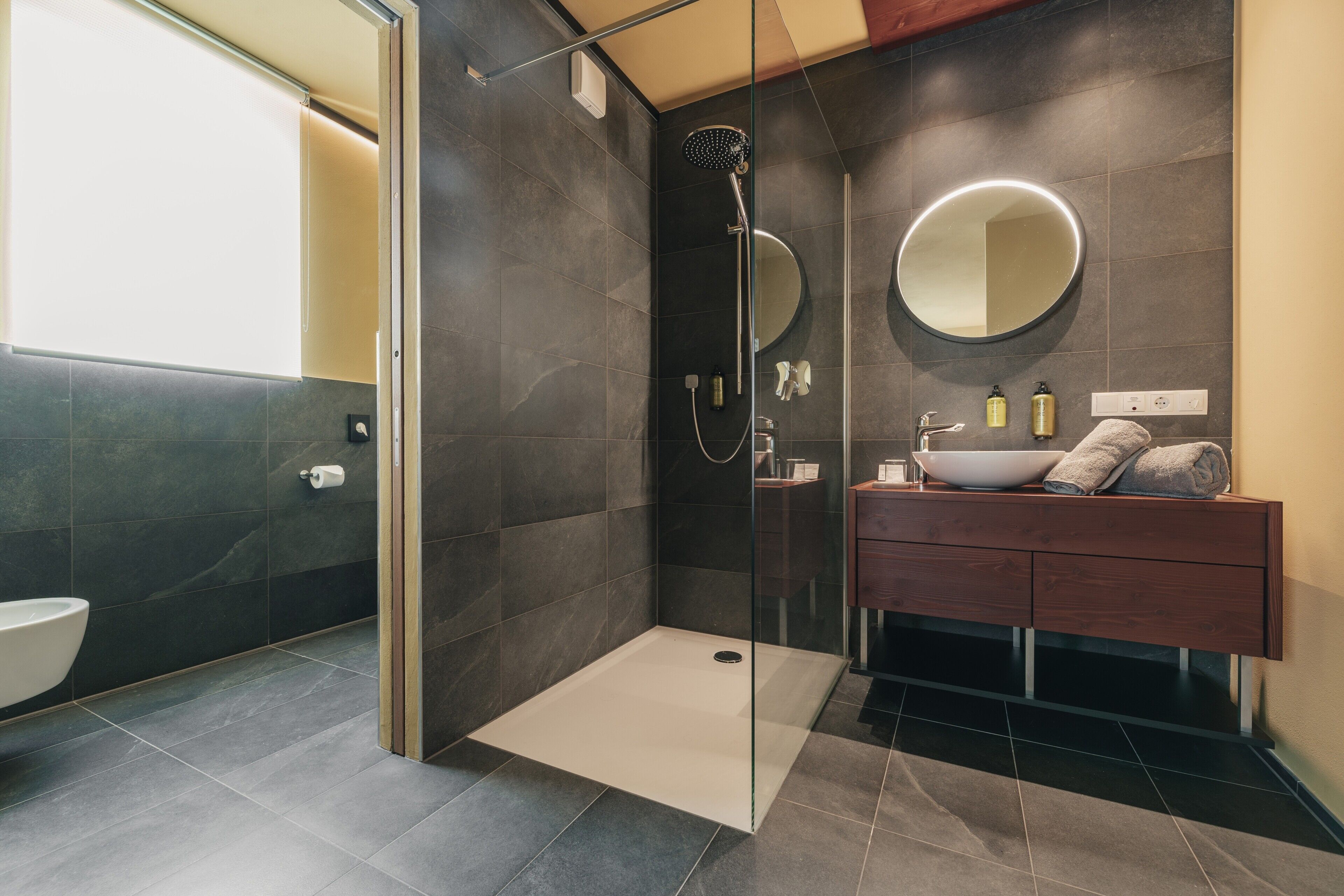 A modern bathroom with a glass-enclosed shower, a round mirror, a wooden vanity, and a toilet.