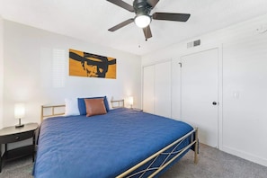 A hotel room with a bed, a ceiling fan, a painting on the wall, and a door.
