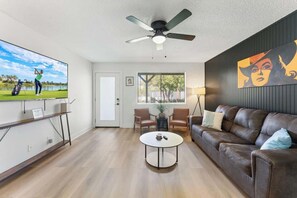 A modern living room with a flat-screen TV, a ceiling fan, a brown leather sofa, a coffee table, and a painting on the wall.