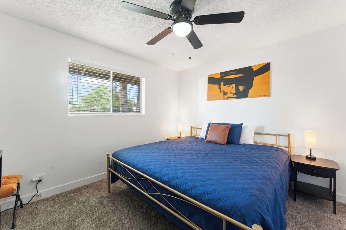 A bedroom with a bed, a ceiling fan, a window with blinds, a nightstand, and a wall-mounted artwork.