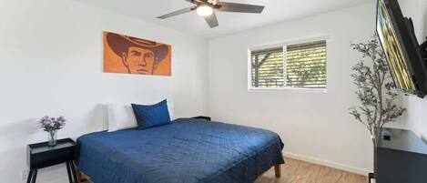 A bedroom with a bed, a ceiling fan, a window with blinds, a nightstand with a vase, and a wall-mounted TV.