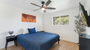 Quarto com cama, ventilador de teto, janela com persianas, mesa de cabeceira com um vaso e televisão fixada na parede.