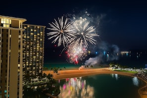 Hotel alto com vista para a praia, piscina refletindo luzes coloridas e área de praia com palmeiras.