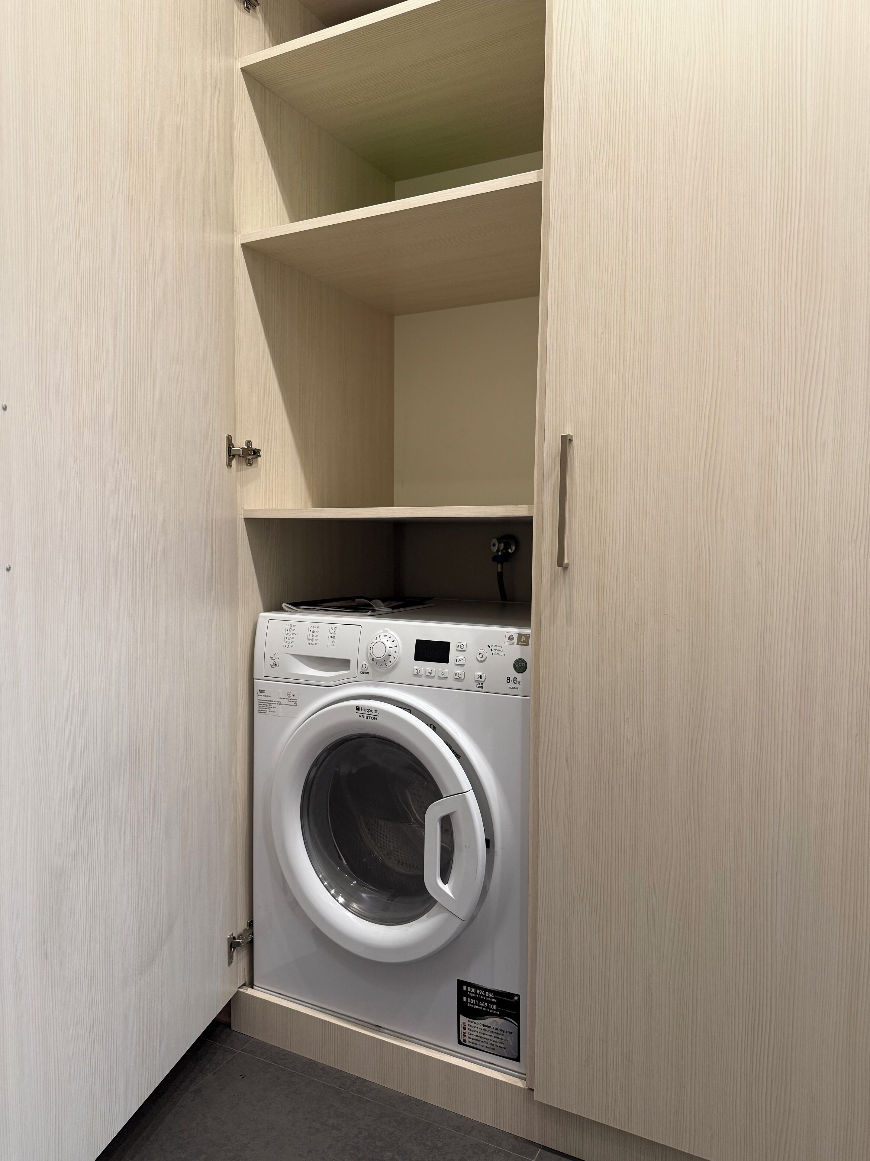 Una lavadora compacta e integrada en un pequeño cuarto de lavado, con armarios de madera y suelo de baldosas.