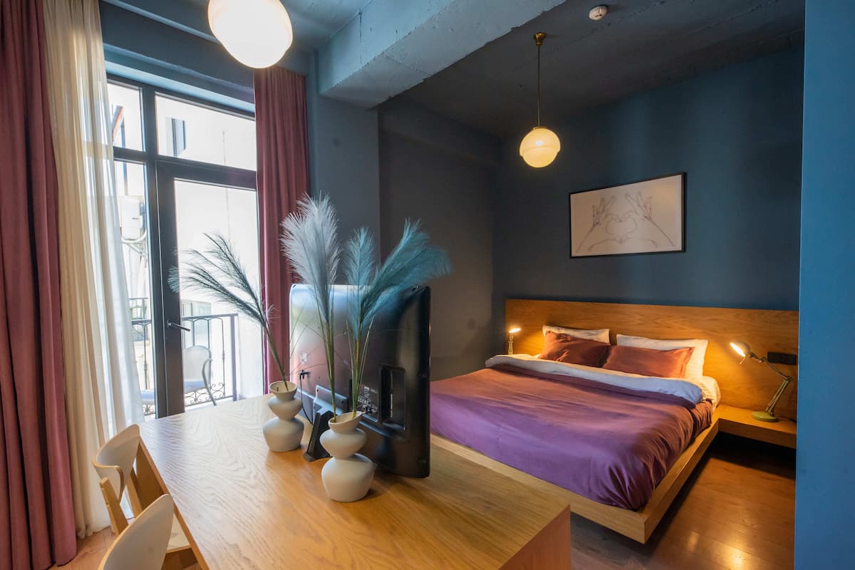 A modern bedroom with a bed, bedside table, lamp, and a view of the outdoors through a window.