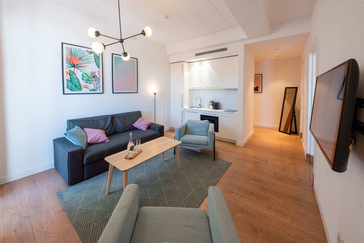 A modern living room with a sofa, a coffee table, and a dining area. There is a kitchen in the background with white cabinets and a sink.