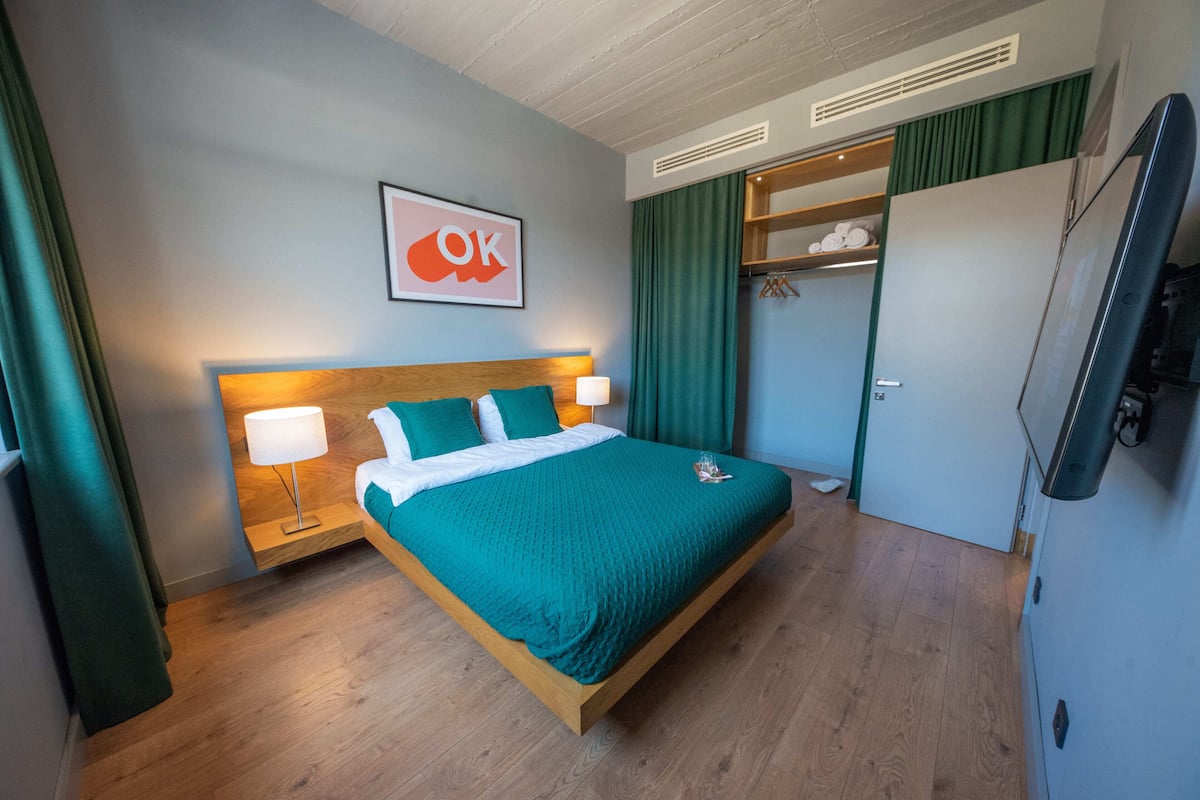 A modern hotel room with a wooden bed, teal bedding, a wall-mounted TV, and a green curtain.
