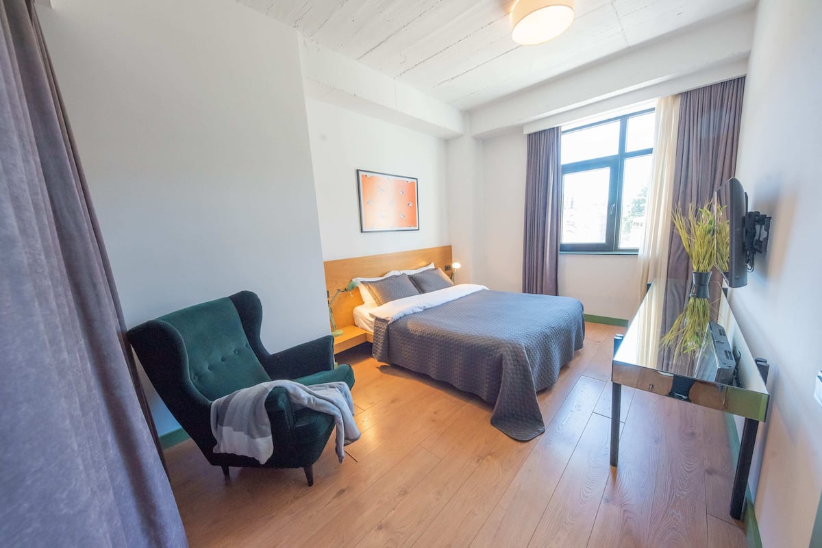 A hotel room with a bed, a chair, a desk with a plant, and a window with curtains.