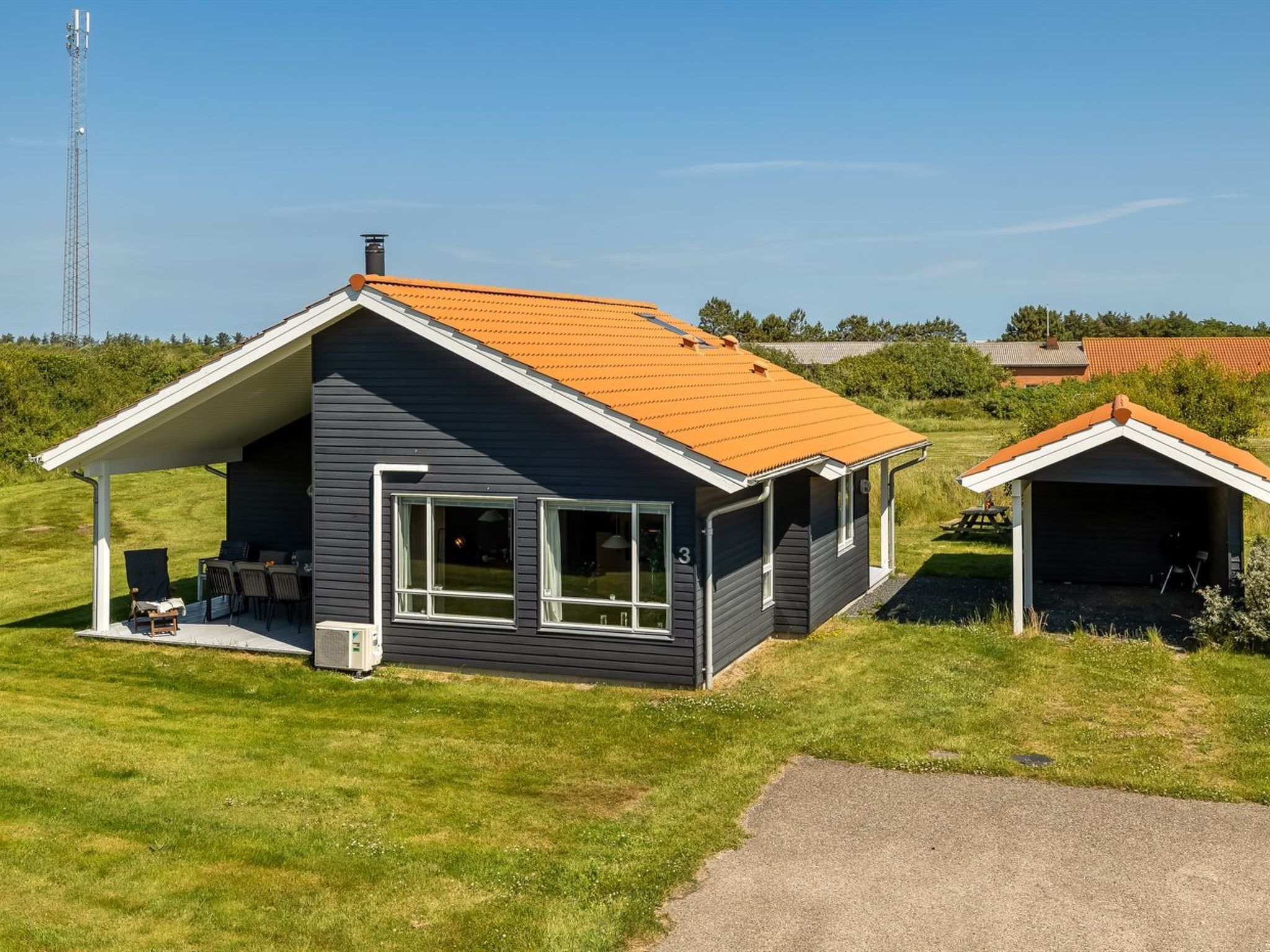 Une maison moderne à deux étages, dotée d’une façade sombre et d’un toit orange, comprenant un patio couvert avec des meubles d’extérieur et un petit abri de jardin.