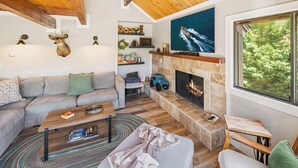 A cozy living room with a stone fireplace, a large TV mounted above it, and a view of greenery through a window.