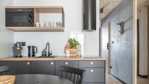 Une cuisine moderne équipée d’un four encastré, d’une machine à café et d’une étagère en bois où sont rangées des tasses et des verres.