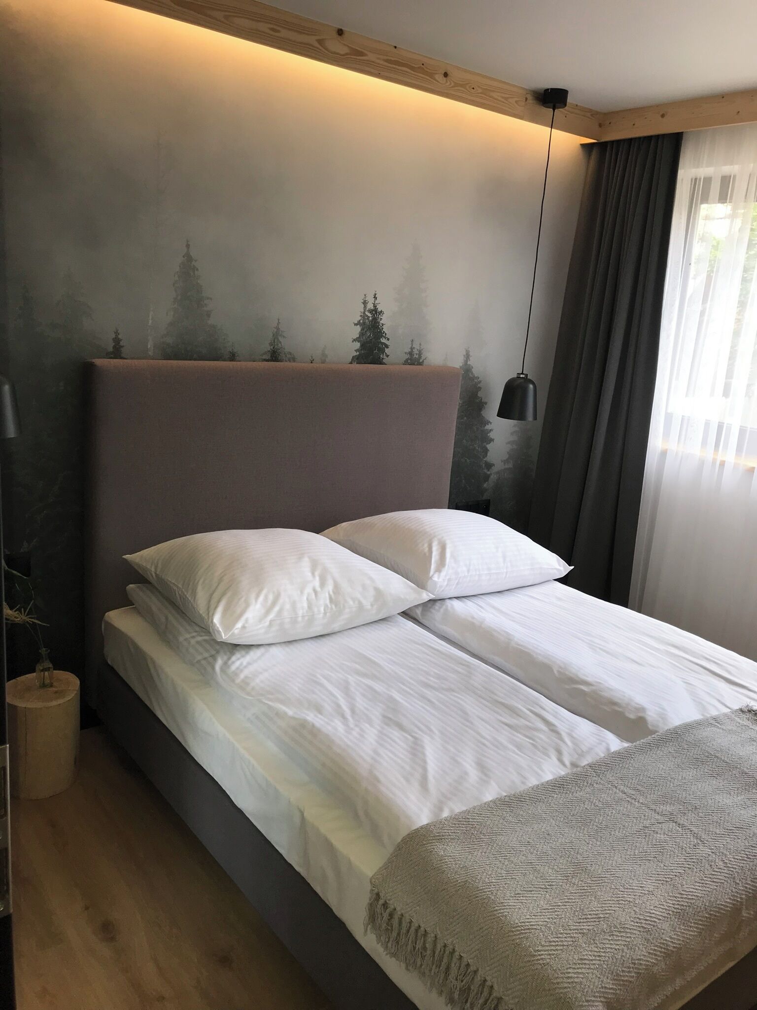 Un lit bien fait, avec du linge de lit blanc et une tête de lit grise, une lampe de chevet et une fresque murale représentant des arbres.