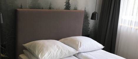 Una cama bien tendida con sábanas blancas y un cabecero gris, una lámpara de noche y un mural de árboles en la pared.