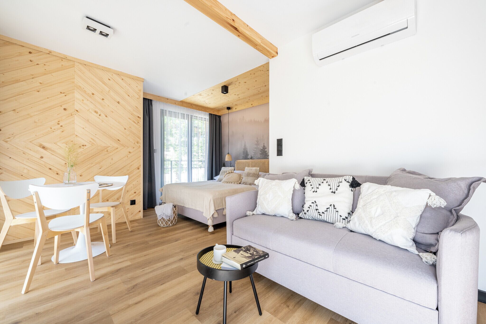 A modern, well-lit room with a wooden floor, a bed with a headboard, a grey sofa, and a dining area with a table and chairs.
