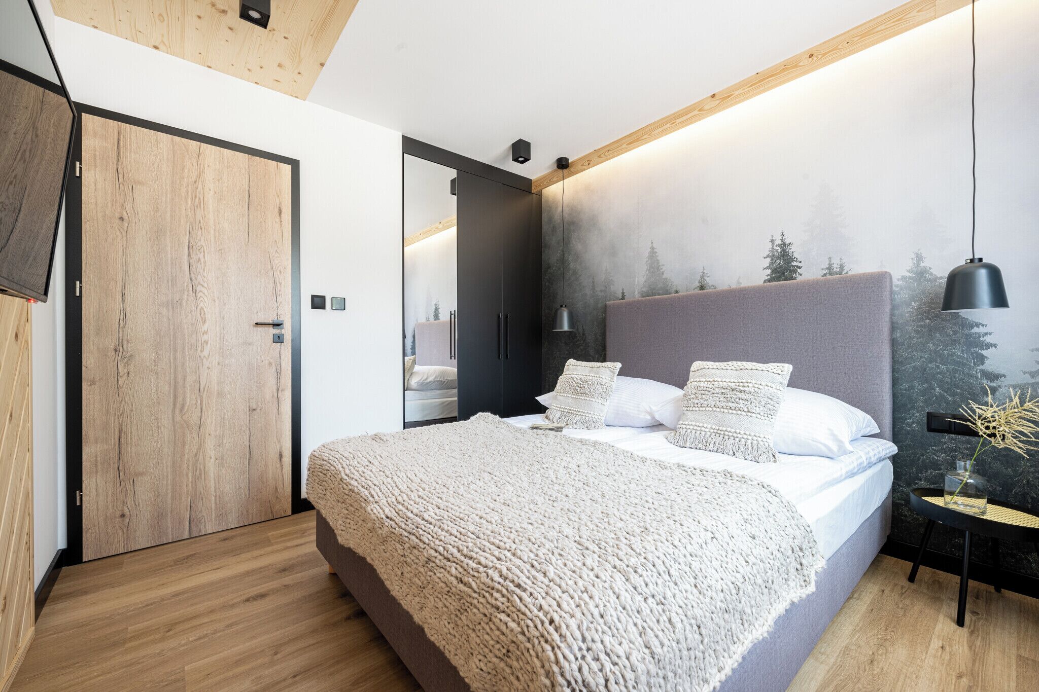 Un dormitorio moderno con una cama grande, una puerta de madera, un televisor montado en la pared y una mesita de noche con una lámpara.