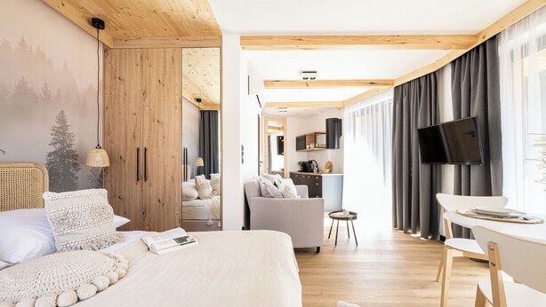 Une chambre d’hôtel moderne avec un plafond en bois, un grand lit recouvert d’une couette blanche, un petit bureau avec une chaise, une télévision et une vue sur la cuisine.