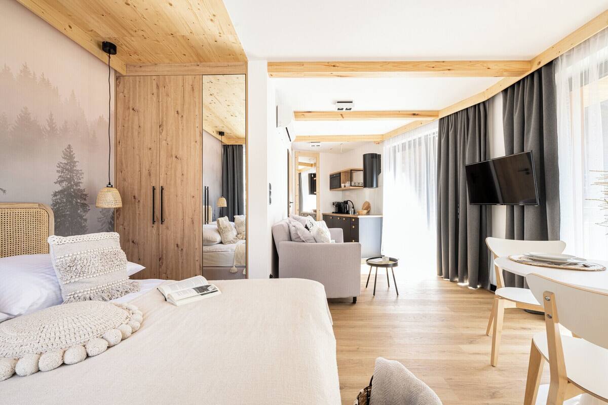 Ein modernes Hotelzimmer mit Holzboden, einem großen Bett mit weißer Decke, einem kleinen Schreibtisch mit Stuhl, einem Fernseher und Blick auf den Küchenbereich.