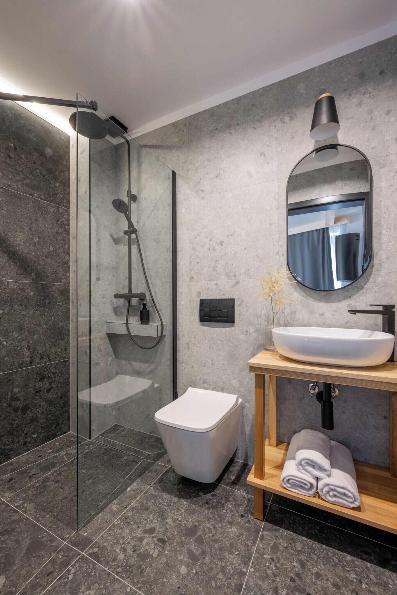 Un baño moderno con ducha de vidrio, una bañera blanca, un espejo redondo y un tocador de madera con lavamanos blanco.