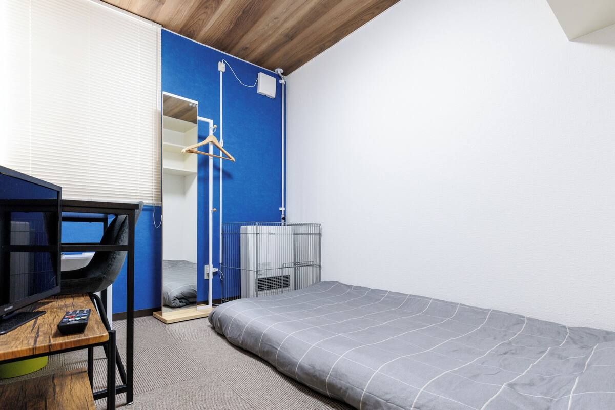 Une pièce comprenant un lit, un bureau avec un ordinateur, une chaise, un porte-manteau mural et une fenêtre avec des stores.