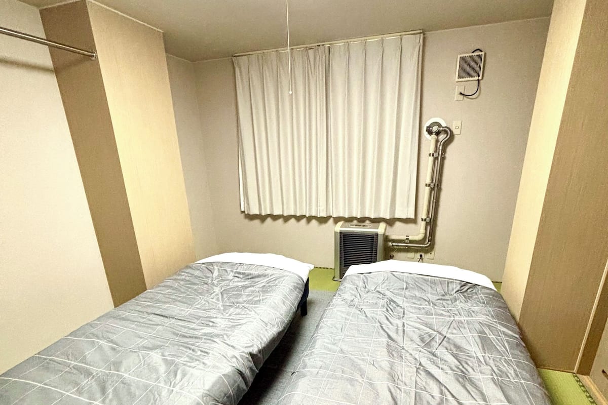 Une petite chambre d’hôtel composée d’une seule pièce, avec deux lits simples, un luminaire au plafond, une fenêtre avec des rideaux et un climatiseur fixé au mur.
