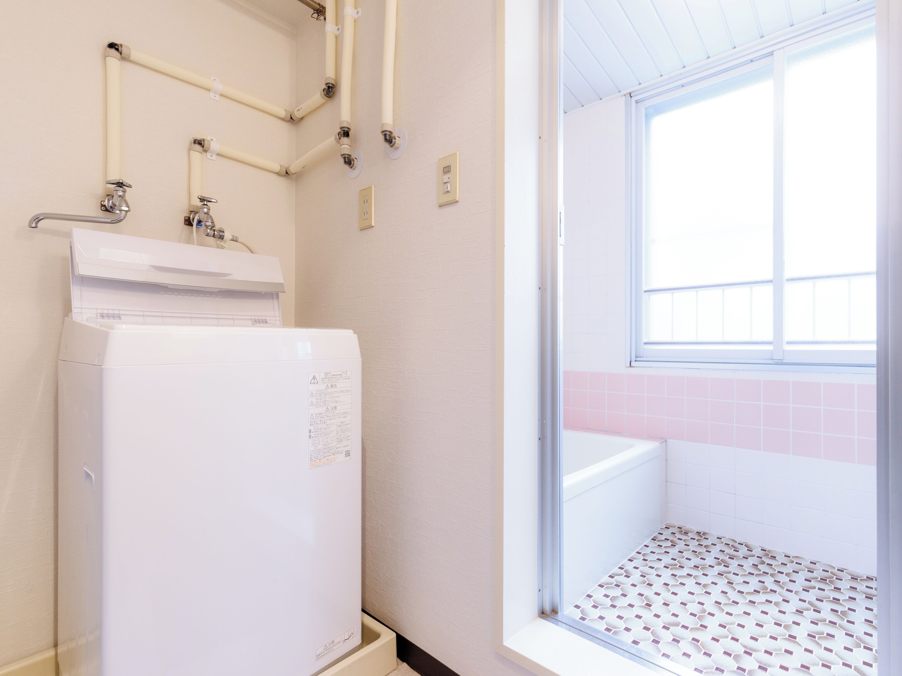 Een badkamer met een witte wasmachine, een raam en roze betegelde muren, en een betegelde vloer.