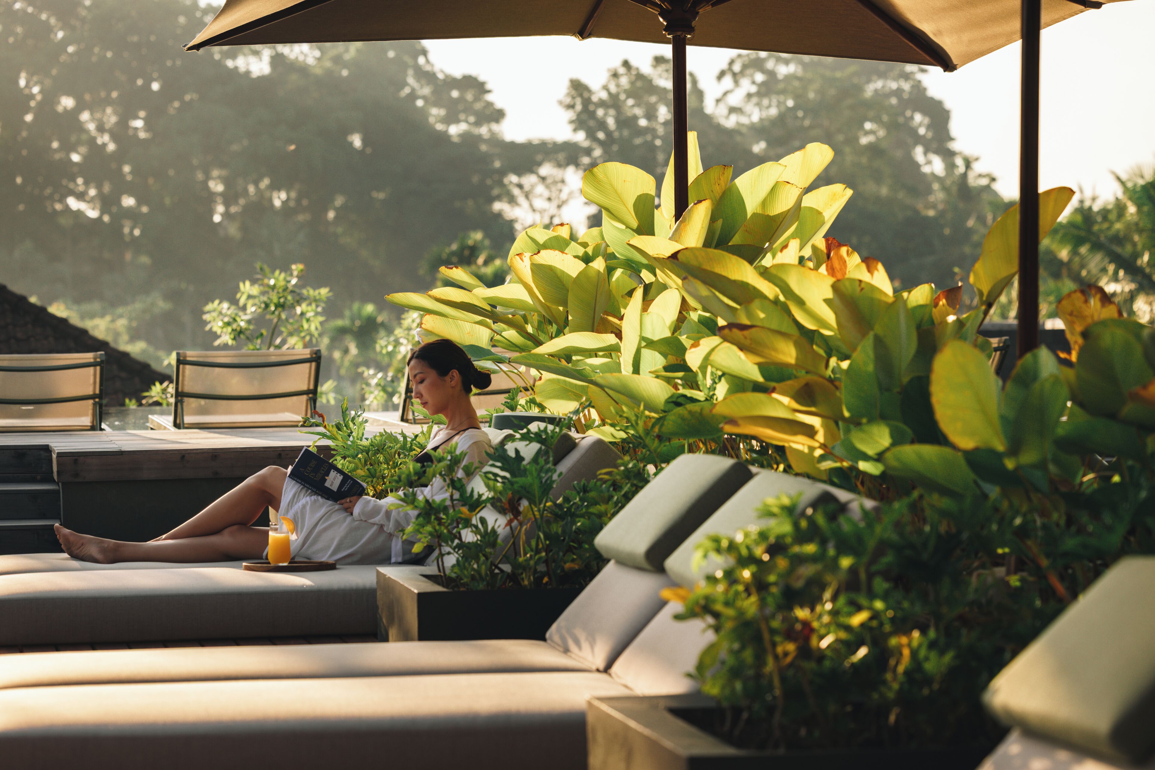 A person relaxing on a cushioned outdoor seating area with a book and a drink, surrounded by greenery and under a large umbrella.