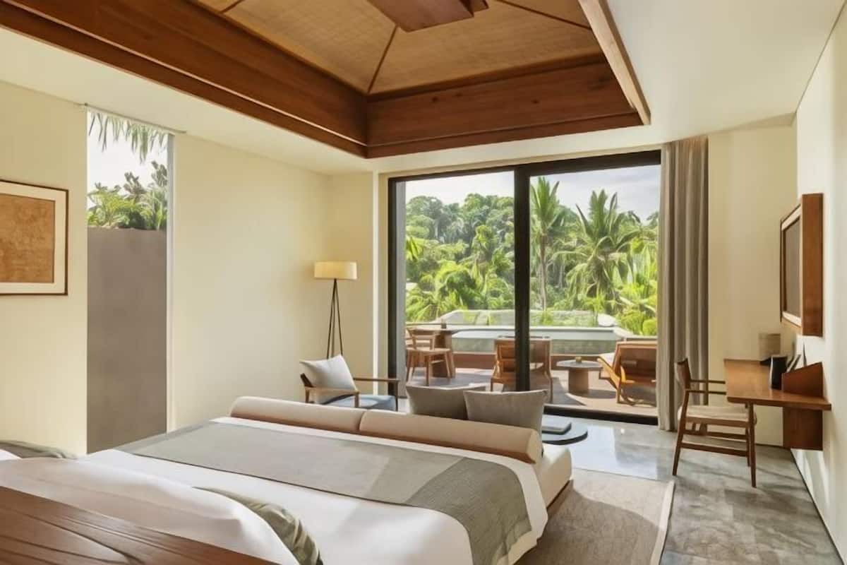 A hotel room with a large bed, a desk, a chair, a lamp, and a view of a lush garden through a large window.