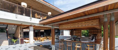 Un moderno espacio al aire libre con una barra de madera, taburetes y una zona de estar con mesas y sillas.