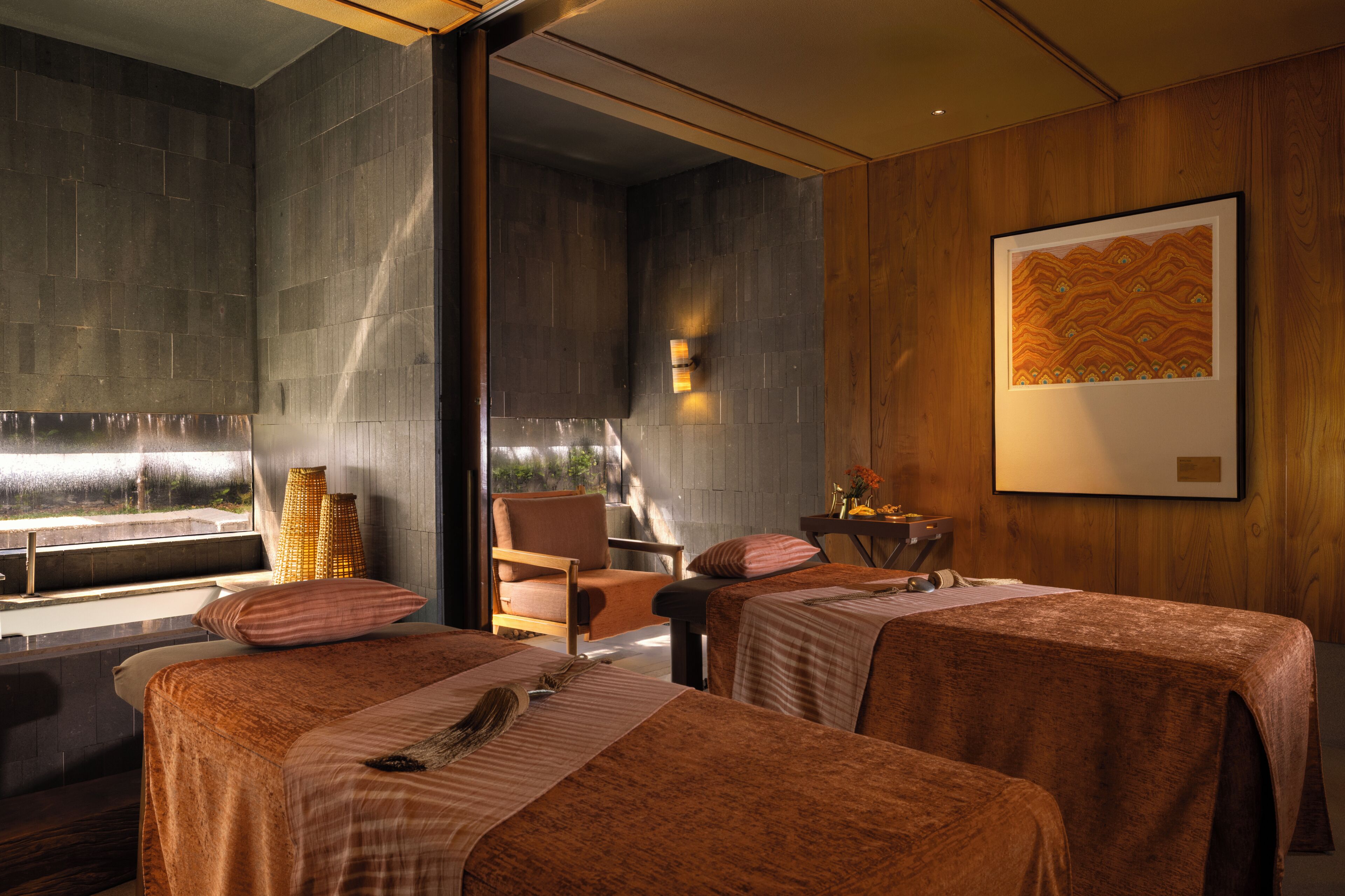 A room with two beds, a wooden chair, a table with a vase, and a framed picture on the wall.