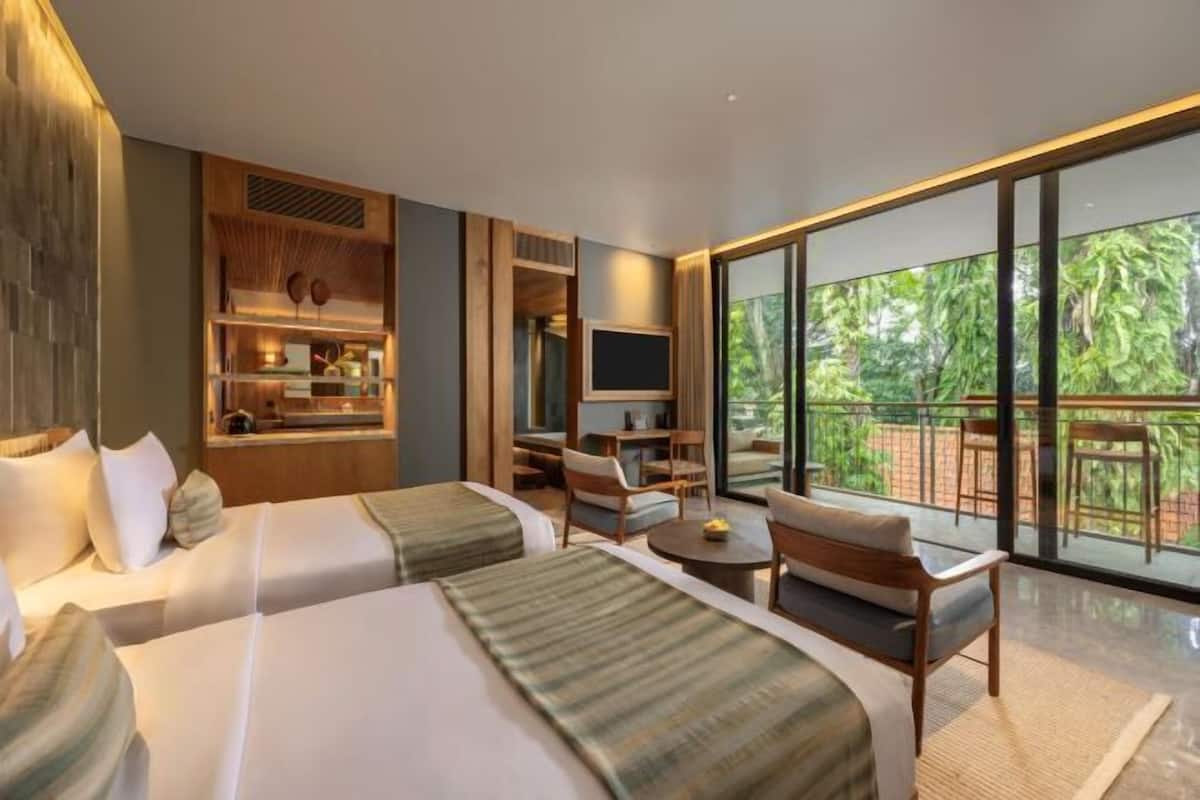 A hotel room with two beds, a TV, a balcony with a view of greenery, and a small dining area.