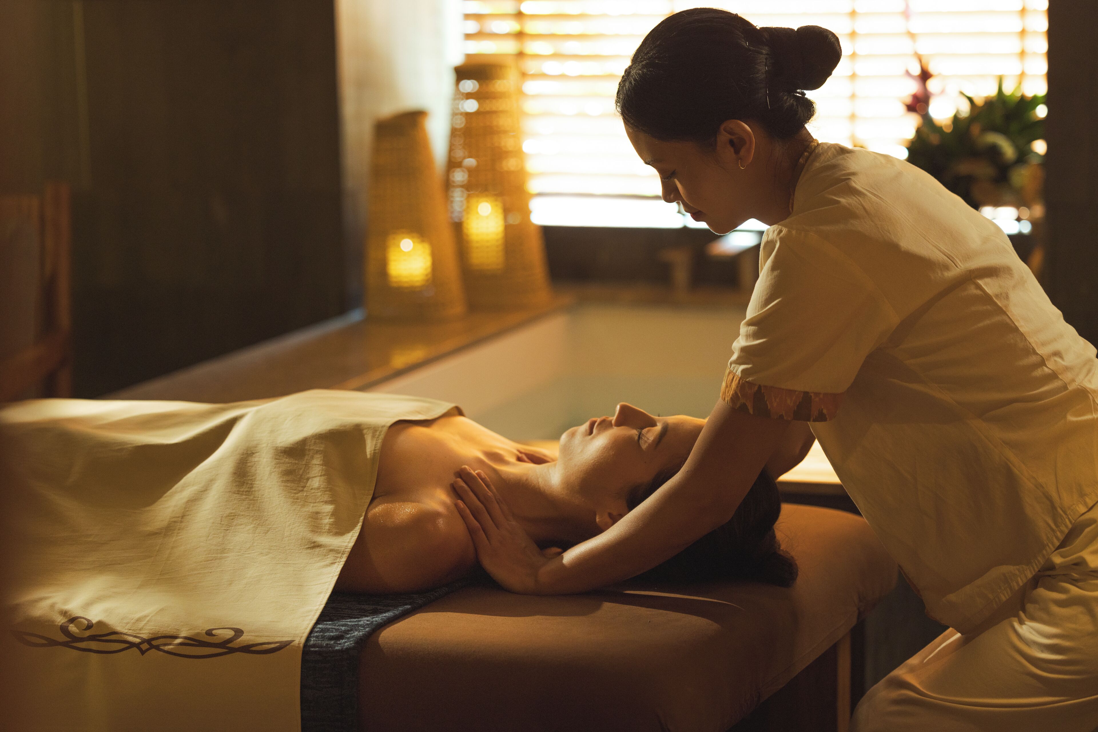 A massage therapy session in a spa setting with a person lying on a massage table, another person performing the massage, and a window with blinds in the background.