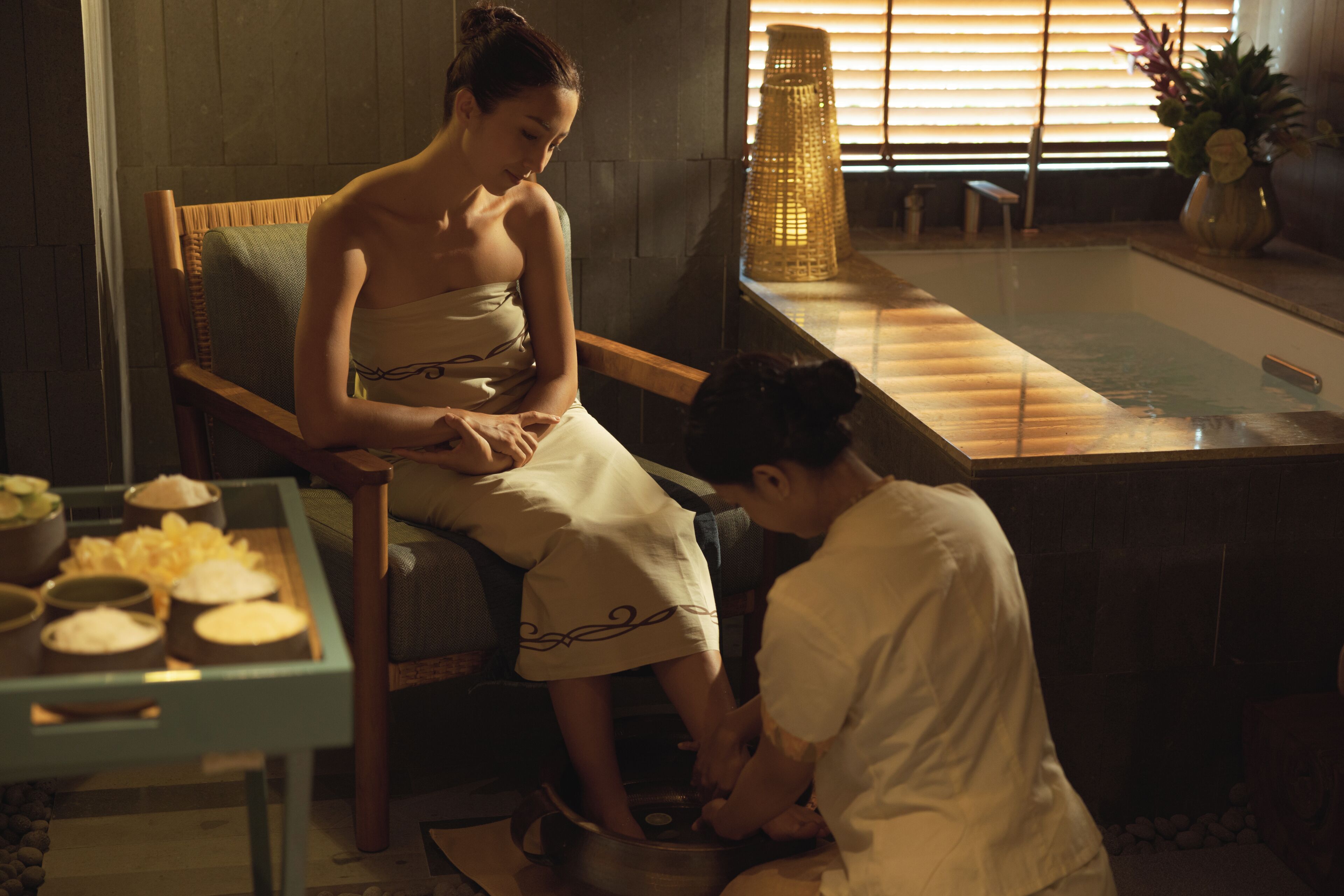 Un ambiente de spa con una silla de madera, una mesa con cuencos y una persona atendiendo a otra sentada.