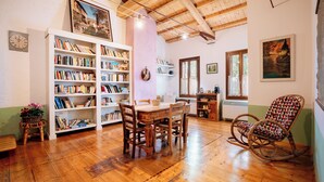 A cozy room with wooden floors, a dining table with chairs, a rocking chair, a bookshelf, and a window with curtains. - Agriturismo Le Noci (Tarzo)
