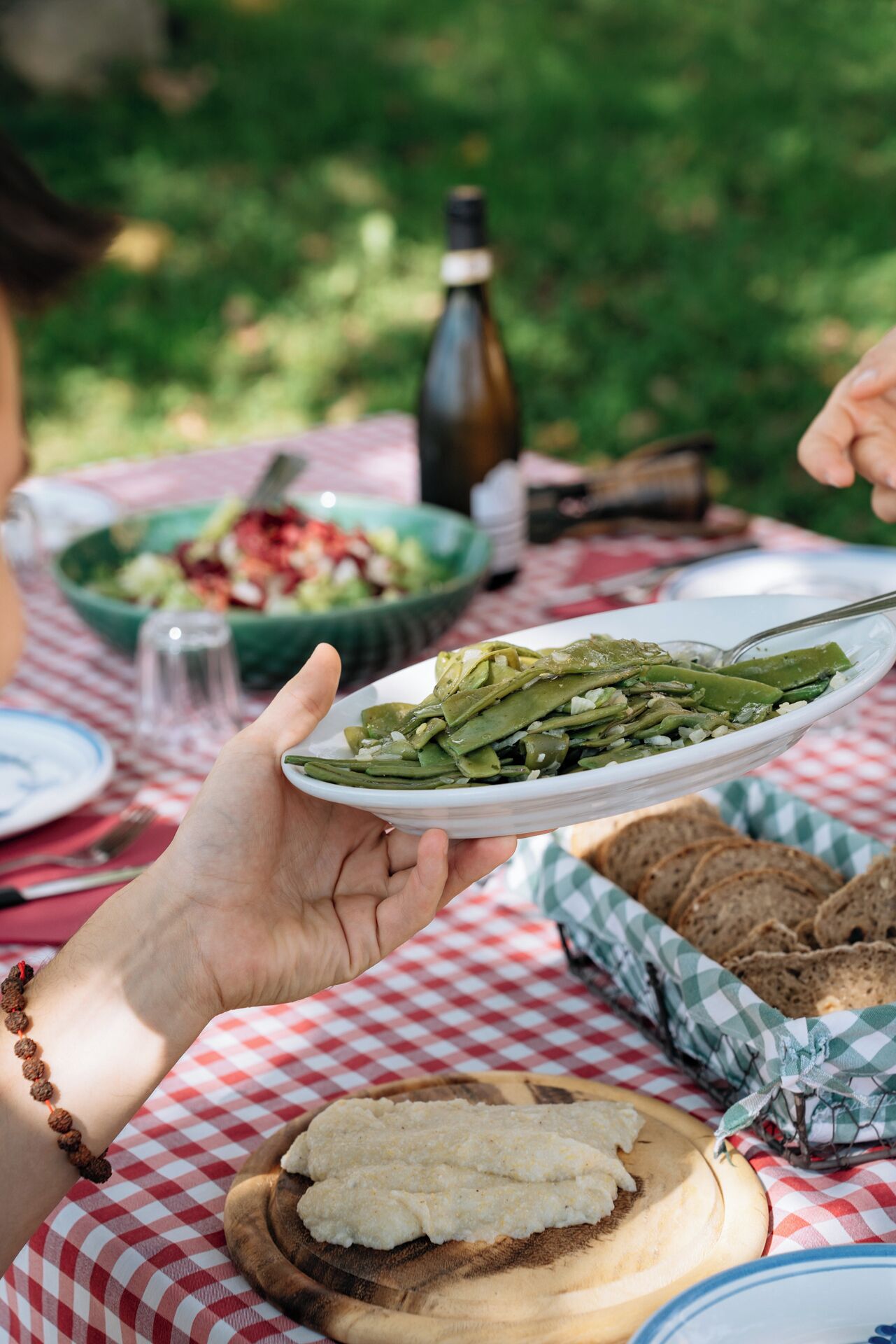 A person holding a plate of green beans at an outdoor dining table with a checkered tablecloth.