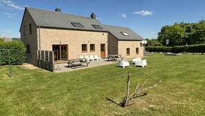 A two-story brick building with a sloped roof, a patio area with outdoor seating, and a well-maintained lawn. - Villa Marie Frances (DURBUY)