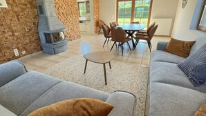 A modern living room with a fireplace, grey sofas, a dining table with chairs, and a kitchen in the background. - Villa Marie Frances (DURBUY)