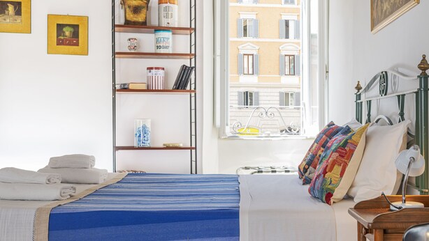 Un dormitorio con cama, ventana con vistas a edificios, estantería y mesita de noche.