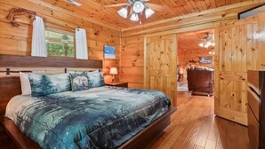 A wooden cabin bedroom with a bed, a ceiling fan, and a view into the living area.