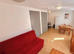 A compact living space with a red sofa, a dining table with chairs, a small kitchen area, and a white refrigerator.