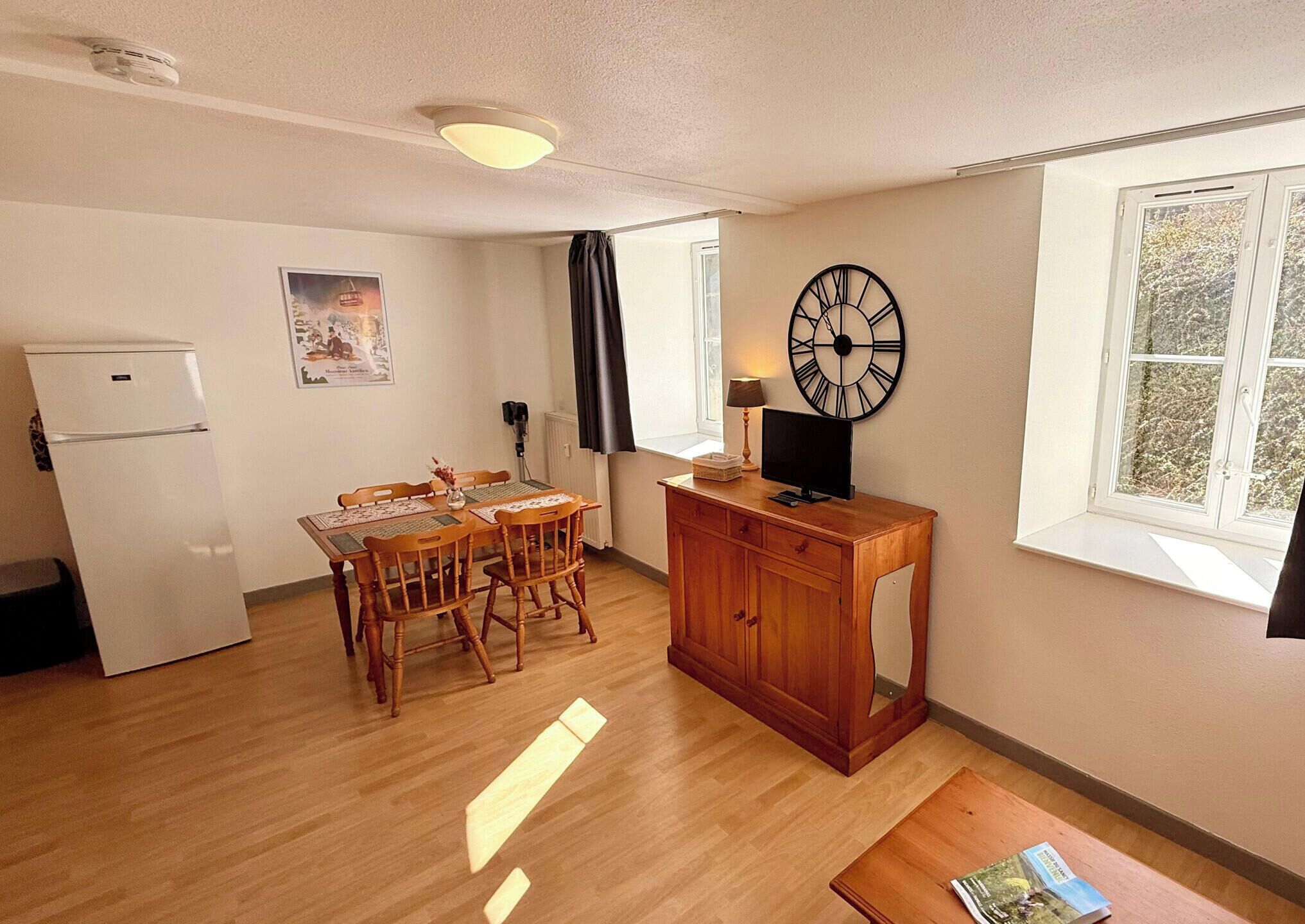 Una cocina compacta con refrigerador, una mesa de comedor de madera, sillas, un pequeño mueble de madera, un reloj de pared y una ventana con vista a árboles.