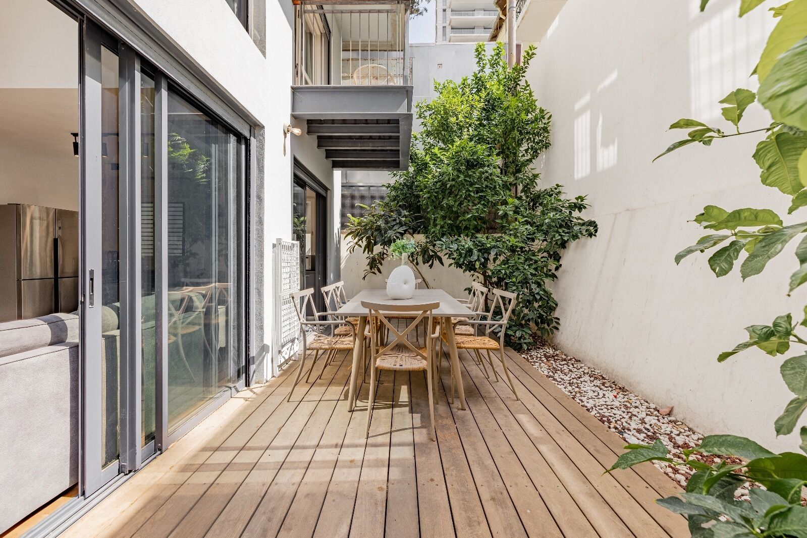A wooden deck with a table and chairs, surrounded by a white wall and a large tree.