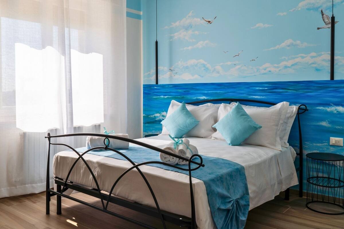 Ein Schlafzimmer mit Bett, einem Fenster mit transparenten Vorhängen, einem Wandgemälde vom Meer und einem kleinen runden Tisch.