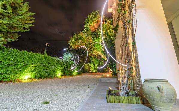 Espaço ao ar livre à noite, com um caminho de cascalho, vegetação e uma instalação luminosa decorativa.