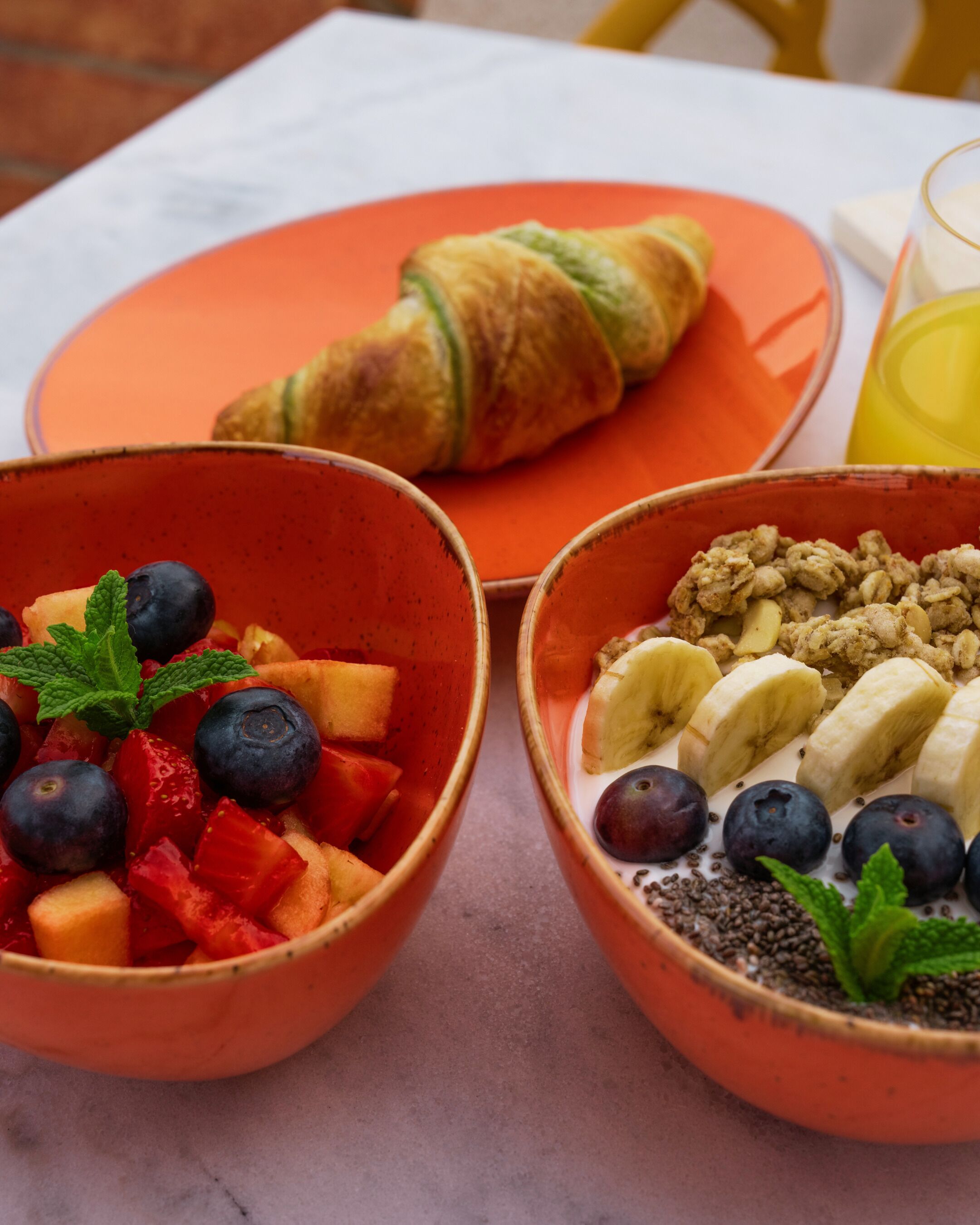 Une table mise pour le petit-déjeuner avec un croissant, deux bols de fruits et de céréales, et un verre de jus d’orange.