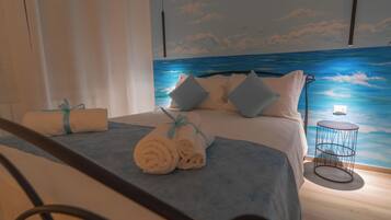 Cama bem arrumada com toalhas enroladas e almofadas, uma mesa lateral redonda e um mural de paisagem marítima na parede.