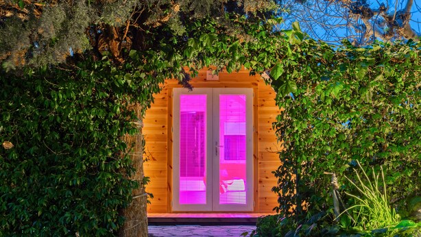 Uma cabana de madeira com entrada iluminada em rosa, rodeada por vegetação.