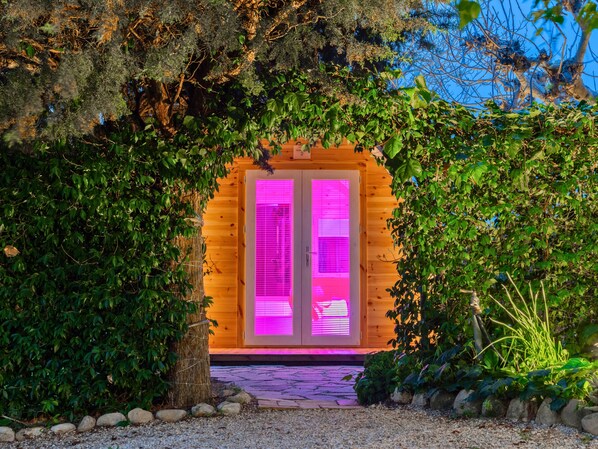 Een houten hutje met een roze verlichte ingang, omringd door groen.