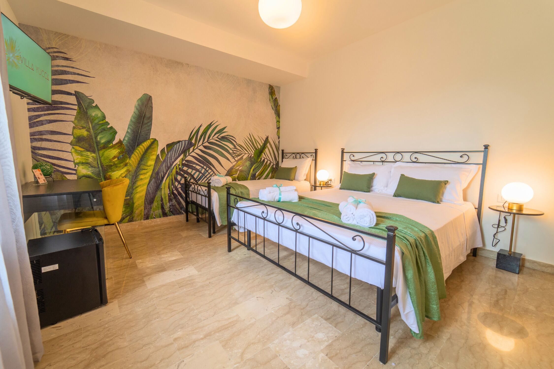 Un dormitorio con cama, mesitas de noche, un escritorio y un mural de hojas tropicales en la pared.