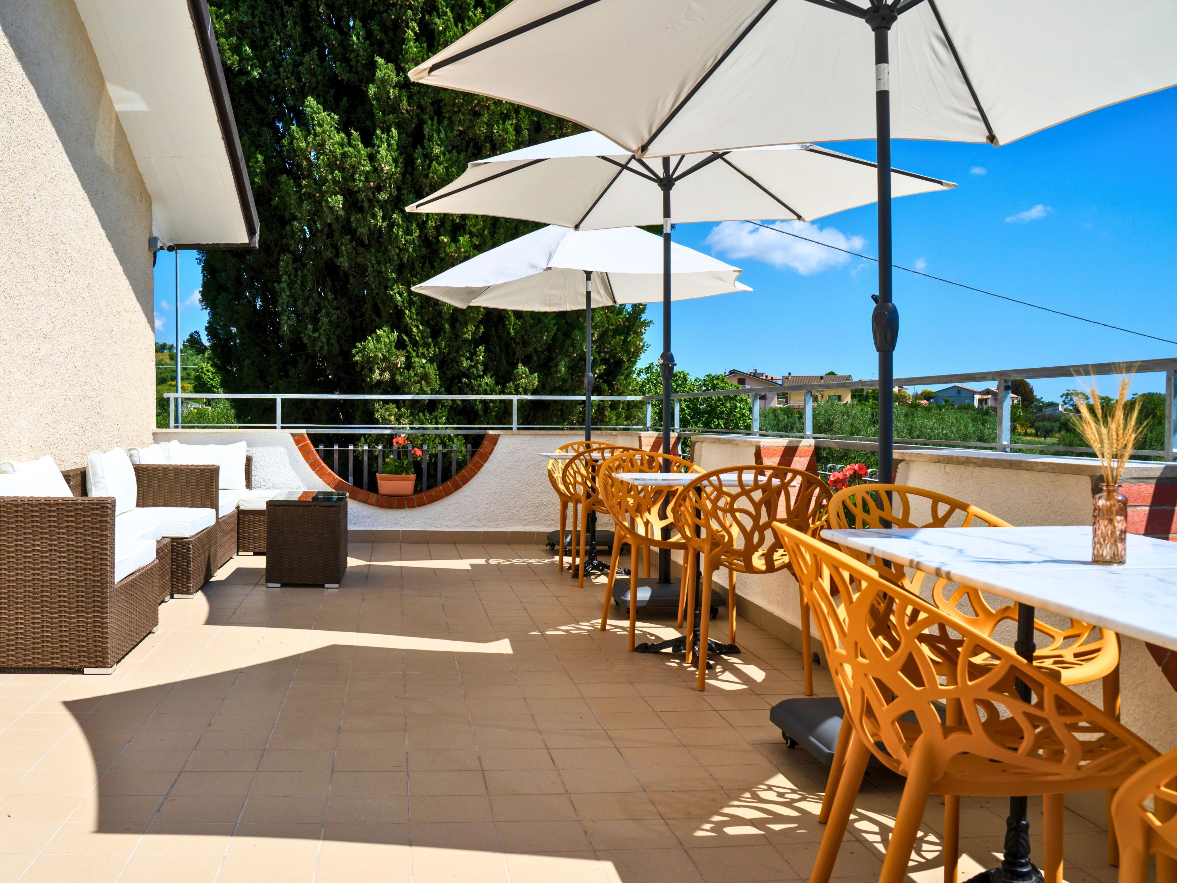 Een terras buiten met rieten meubilair, witte parasols en een uitzicht op groen en gebouwen op de achtergrond.